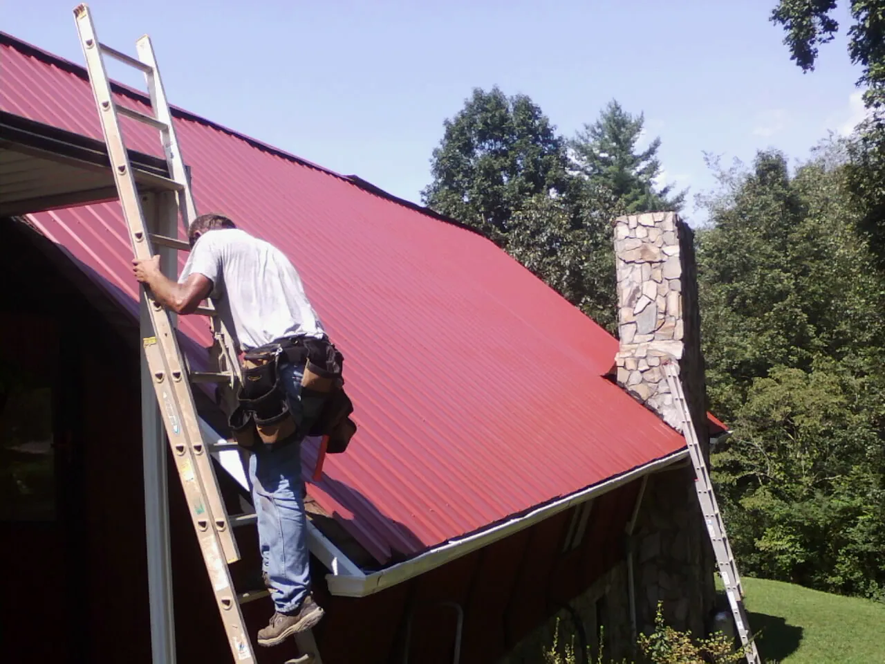 Roofer on a ladder beside a red metal roof and stone chimney — Soffit & Fascia Repair by Roofing Brothers in Manvel